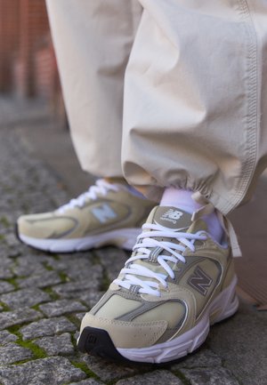 Beżowe buty do biegania New Balance z białymi sznurowadłami, noszone z beżowymi spodniami, stojące na brukowanej nawierzchni.