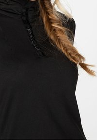 Närbild på en person som bär en svart topp med dragkedja och en ensam blond fläta som vilar på axeln.