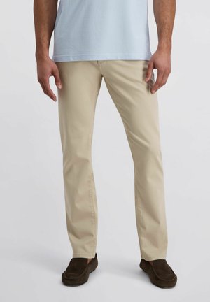 Homme portant un pantalon beige à coupe droite, une chemise bleu clair et des mocassins marron foncé, debout devant un fond clair uni.