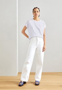 Striped white short-sleeve top with a subtle graphic, paired with high-waisted white jeans and blue platform sandals on a wooden floor.