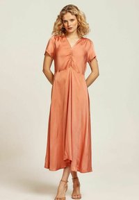 Lång, V-ringad klänning i rostfärg med glänsande tyg, korta ärmar och frontveck, som når till vadknät. Har en flödande silhuett.