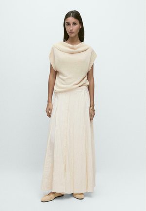 Femme, de face, portant un pull sans manches beige à col bénitier, une longue jupe plissée crème, des ballerines beiges et des bracelets dorés au poignet droit.