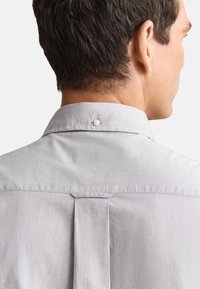 Homme vu de dos portant une chemise gris clair à col avec un pli au dos et un seul bouton sur la patte de col.