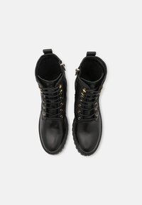 Un paio di stivali da combattimento neri in pelle con lacci, occhielli dorati e cerniere laterali, visti dall'alto su uno sfondo bianco.