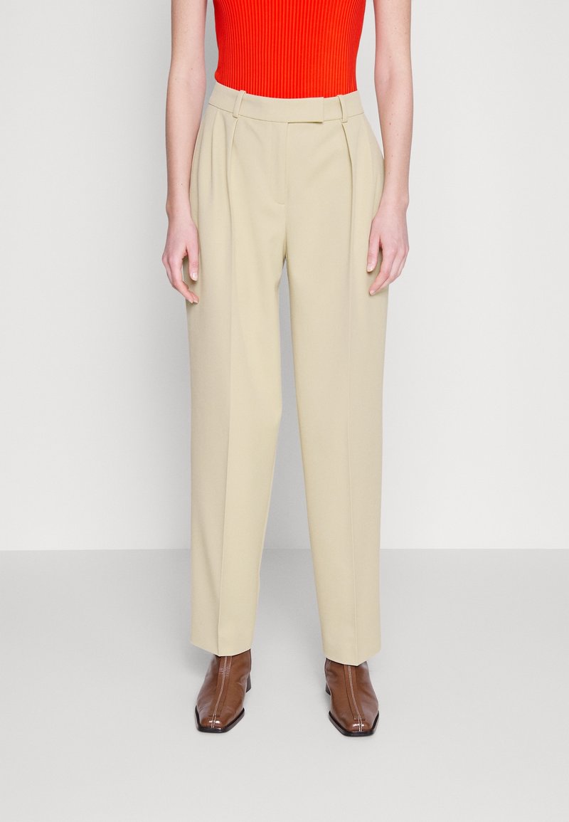 Femme portant un pantalon beige taille haute à jambes larges, un haut rouge sans manches, et des bottines marron à bout carré, debout devant un fond uni.