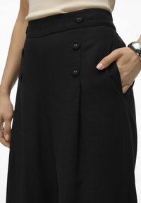 Pantalons noirs taille haute avec une fermeture à boutons, des plis et des poches latérales, confectionnés dans un tissu lisse au toucher doux.