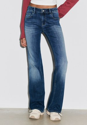 Kvinde iført blå vidde jeans med høj talje og falmede detaljer, hvide sneakers og en hvid crop top med lange røde ærmer.