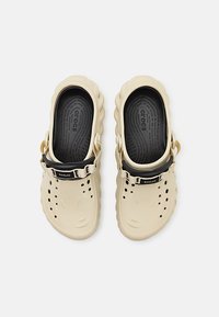 Sabots Crocs beiges avec semelles intérieures noires et sangles réglables, perforés pour la ventilation, montrés en vue de dessus sur un fond blanc.