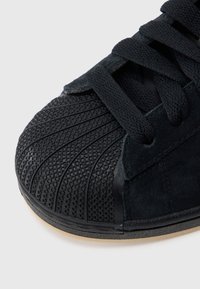 Μαύρο αθλητικό παπούτσι με υφασμένη λαστιχένια μύτη, πάνω μέρος από σουέτ, επίπεδα μαύρα κορδόνια και λεία σόλα. Διαθέτει διαγώνια και ρόμβου σχέδια στη μύτη.