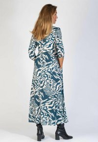 Długa, turkusowa sukienka z białymi abstrakcyjnymi wzorami, z rękawami do trzech czwartej i bocznymi kieszeniami, wykonana z lekkiego materiału. Czarne buty akcentują całość.
