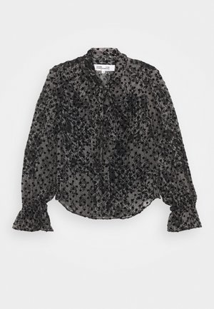 Schwarze transparente Langarmbluse mit Blumen- und Polka-Dot-Mustern, Rüschenmanschetten und einem Bindeband am Hals auf hellem Hintergrund.