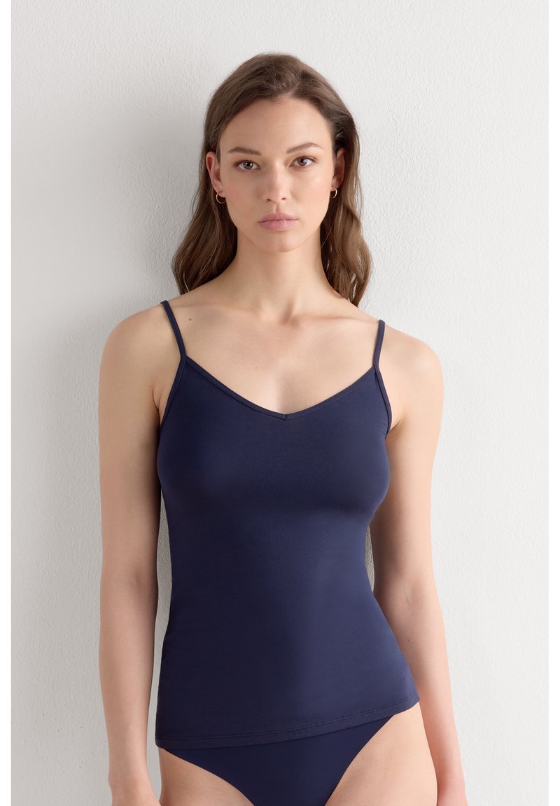Junge Frau mit langen braunen Haaren trägt ein marineblaues Camisole und passende Unterteile, steht vor einer schlichten hellen Wand und schaut nach vorne.