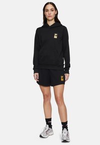 Svart huvtröja med guldlogga, avslappnad passform, framficka, i kombination med svarta shorts, silverfärgade sneakers och svarta strumpor med logga.