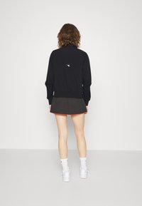 Svart jacka med uppvikta ärmar, med en liten vit logotyp på baksidan. Bärs över en svart kjol med röd kant. Vita strumpor och skor.