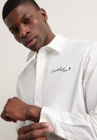 Camisa blanca con cuello clásico, que presenta un detalle de firma bordada en negro. Tejido suave con puños abotonados, ajuste entallado.