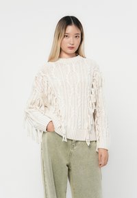 Cremfarbener Kabelstrickpullover mit Fransenakzenten an den Ärmeln, runder Ausschnitt und gerippten Bündchen, kombiniert mit leichten, hellgrünen weit geschnittenen Hosen.