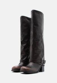 Ζευγάρι σκούρες καφέ δερμάτινες μπότες μέχρι το γόνατο με χοντρούς τακούνια και λουράκια στον αστράγαλο με ασημένιες αγκράφες σε λευκό φόντο.