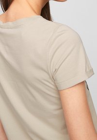 Beige tričko s krátkým rukávem a kulatým výstřihem, s ohrnutými manžetami, měkkou texturou a minimalistickým designem, částečně viditelné ze zad.