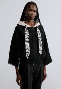 Sudadera negra con cremallera y capucha de color crema, con cordones a rayas de cebra y mangas anchas. El material parece suave con una textura lisa.