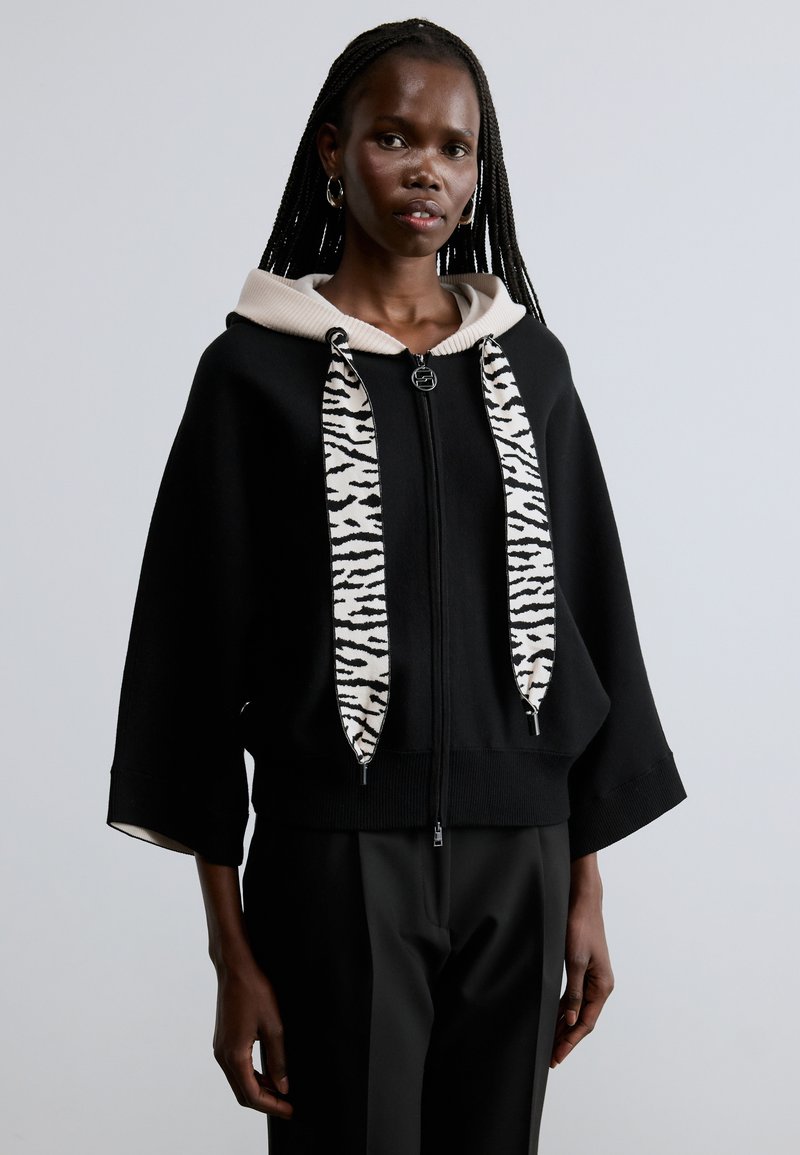 Sudadera negra con cremallera y capucha de color crema, con cordones a rayas de cebra y mangas anchas. El material parece suave con una textura lisa.