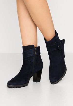Femme portant des bottines en daim bleu marine avec talons blocs, fermeture éclair latérale et bride décorative avec boucle sur fond clair.