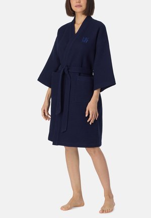 Femme portant un peignoir bleu marine à la texture gaufrée, longueur genou, avec des manches larges, deux poches avant et une ceinture nouée.