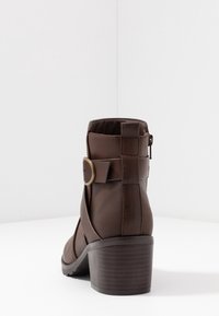 Botín marrón hasta el tobillo con punta redonda, cuero texturizado, cremallera lateral y un pequeño tacón de madera. Presenta una correa y un detalle de hebilla de latón.