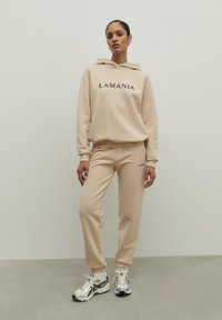 Beige mikina s kapucňou a zodpovedajúce jogger nohavice s textom "LAMANIA", vyrobené z mäkkej tkaniny, s manžetami na členkoch a bočnými vreckami.