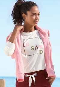 Helle rosa Zip-Hoodie über einem weißen T-Shirt mit buntem Text. Kastanienbraune Jogginghose mit metallischen Ösen, glatter Stoff, lässige Passform.