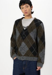 Cardigan con motivo argyle marrone, grigio e nero, con scollatura a V, chiusura con bottoni e tessuto di lana morbido e testurizzato. Indossato sopra una camicia chiara.