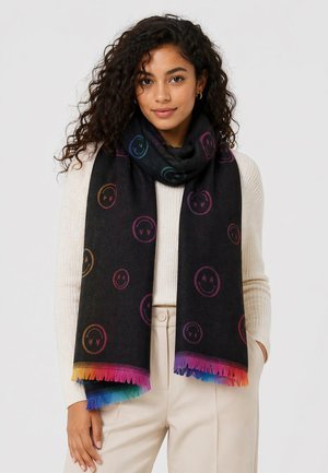 Young woman wearing a black scarf with colorful smiley faces and rainbow fringe, cream sweater, and beige pants, standing against a white background.