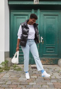 Svart pufferväst över en vit långärmad tröja, i kombination med ljusblå jeans och vita sneakers. Håller en liten vit handväska. Grön dörr i bakgrunden.