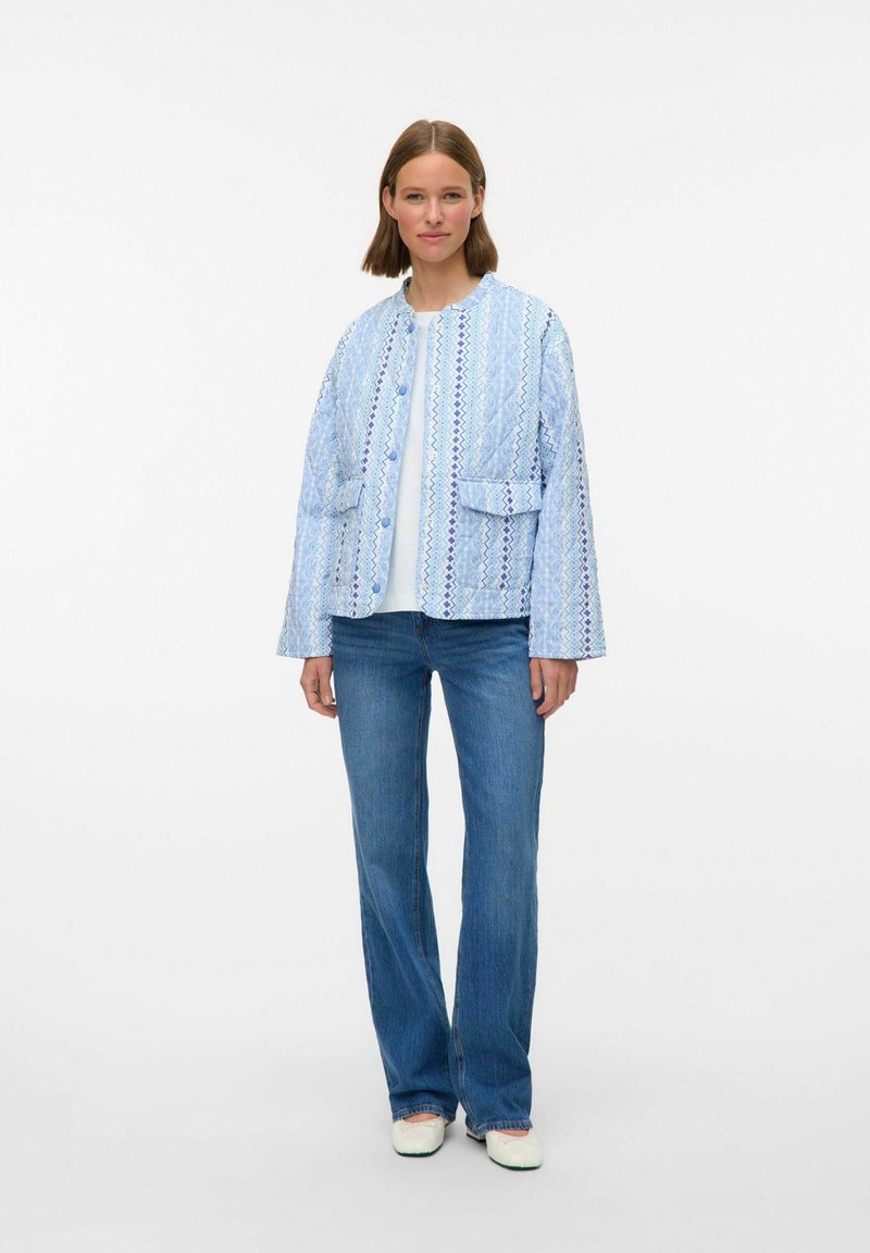 Ljusblå vadderad jacka med mönstrad textur, framknappar och två fickor, i kombination med blå jeans och krämfärgade skor.