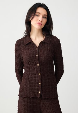 Femme aux cheveux bruns portant une chemise marron texturée boutonnée et un pantalon assorti, sur un fond clair uni.