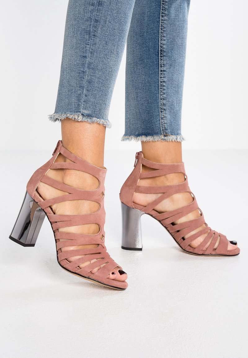 Pieds dans des sandales à talons hauts strass en daim rose avec des talons en bloc métalliques, associées à un jean en denim bleu cropped avec des ourlets effilochés.