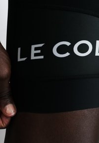Lähikuva tumman ihonvärisestä kädestä, joka lepää mustien shortsejen lähellä, joissa on valkoinen "LE COQ" teksti reisialueella.