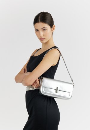 Giovane donna con capelli scuri in un abito nero senza maniche, in piedi con le braccia incrociate, che porta una borsa argento a tracolla su uno sfondo semplice.