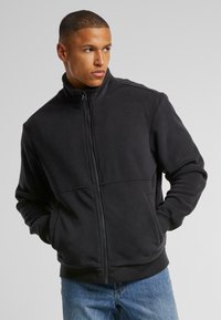 Must musta tõmblukuga fleece-jakk kõrge kraega ja küljetaskutega. Omab sujuvat tekstuuri ja istuvat disaini, koos helesiniste teksadega.