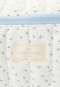 Quiltet stoff i hvitt med små blå og grønne blomstermønstre. Har en lys blå glidelås og en kremfarget merkelapp.