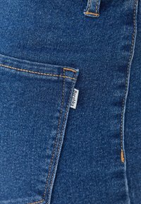 Denim fabric in deep blue with visible stitching in orange, featuring a front pocket and a label stitched on the side.