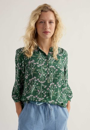 Jonge vrouw met blond golvend haar, gekleed in een groene bloes met bloemenprint en lichtblauwe broek, met handen in de zakken, neutrale achtergrond.