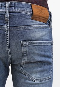 Närbild av en bakficka på blå denimjeans med synlig sömnad och en brun läderetikett som står "Jack & Jones Jeans Intelligence."