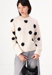 Krämsweter med svarta prickar, stickad textur, ribbad halsringning och ärmslut, parat med grå jeans och en svart sammetväska med kedjeslinga.