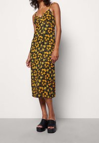 Svart ärmlös klänning med gult och grönt blommönster, knä-lång. Buren med svarta platåskor som visar en slät textur.