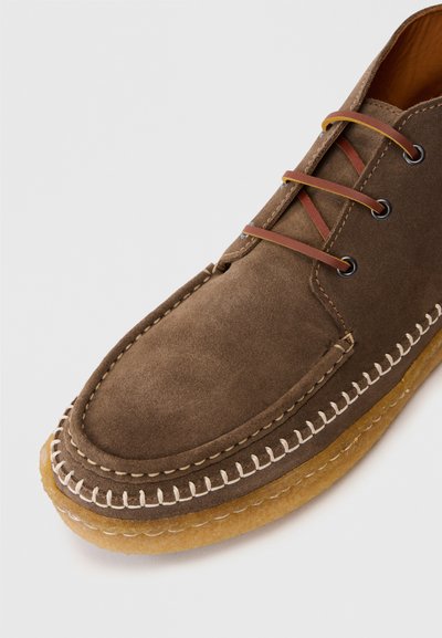 Chaussure en daim marron avec un bout arrondi, coutures beiges et semelle en caoutchouc tan. Équipée de lacets marron et d'œillets en métal pour un maintien sûr.