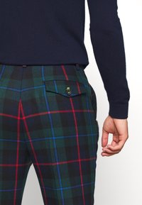 Spodnie w zielono-czarną-czerwoną kratę; wykonane z materiału o gładkiej teksturze; wyposażone w kieszenie zapinane na guziki.