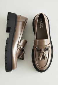 Bruna läderslippers med en svart kraftig sula, med dekorativ kedja och tofsdetalj på ovansidan. Slät yta och rund tå.