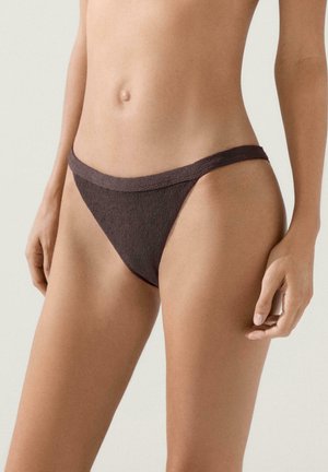 Donna che indossa slip bikini testurizzati marrone scuro a vita alta, in piedi con le braccia rilassate contro uno sfondo chiaro e semplice.