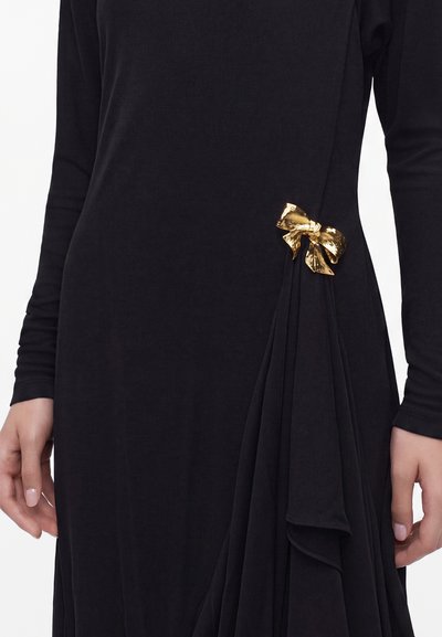 Robe noire à manches longues avec un détail de tissu froncé et une petite broche en nœud doré brillant sur le côté, près de la taille.