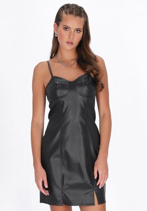 Ung kvinde med flettet hår iført en sort ærmeløs kjole i imiteret læder med tynde stropper og kædehalskæde mod en hvid baggrund.